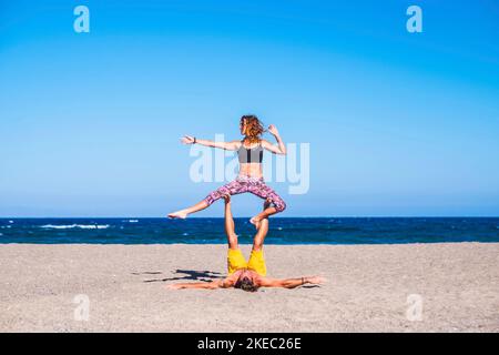 couple of adults doing exercise at the beach togetehr on the sand - two people doing yoga or acro yoga with the sea at the background - man holdinng his girlfriend with his legs Stock Photo