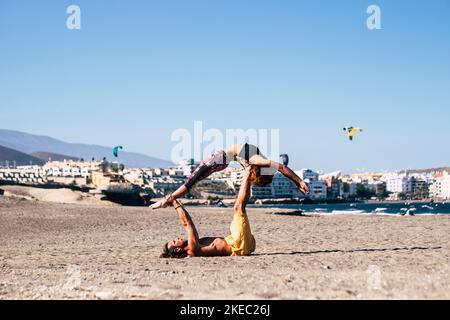 couple of two fitness and active people doing exxercise together in the beach on the sand - adults woman and man doing acroyoga to be healthy Stock Photo