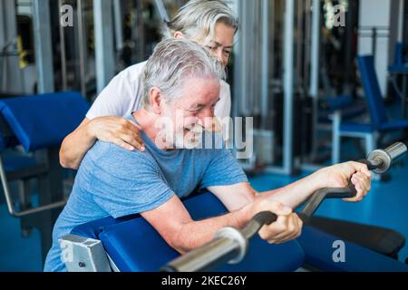 couple of seniors at the gym doing exercise together - mature woman looking and helping her husband holdinng a barr and lifting weight Stock Photo