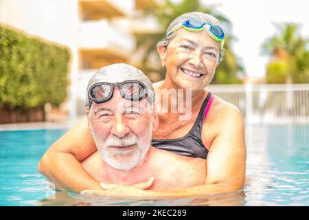 couple of seniors enjoying summer and having fun together in the pool swimming and smiling looking at the camera - cheerful happy people in their vacations - portrait of two mature and retired pesioners Stock Photo