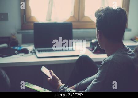 one young man using phone and looking at the laptop or computer at home in his room playing video games. Teenager addicted at social network and social media Stock Photo