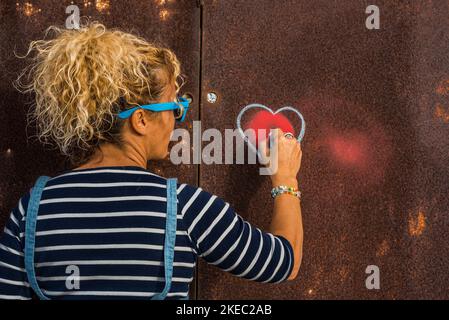 one woman painting a heart on a wall in the street - curly woman showing her emotions Stock Photo
