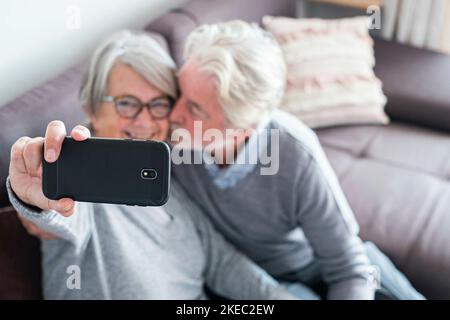 cute couple of two seniors married taking a selfie together sitting on the sofa at home - mature man kissing his wife while she's taking a photo Stock Photo