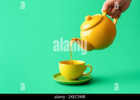 Woman's hand pour tea from yellow jug in a cup, minimalist on a green table. Hot healthy drink, mint tea in a yellow-colored cup. Stock Photo