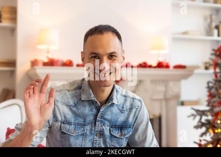 Head shot web camera view happy young handsome man with decorated festive tree on background holding video call, greeting with Happy New year or Merry Stock Photo