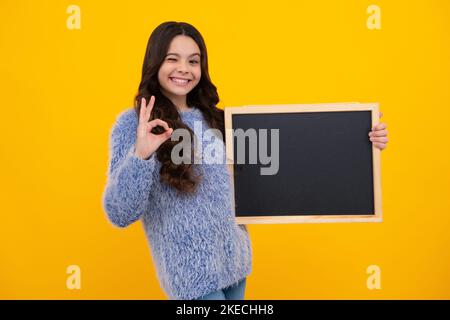 Teenager younf school girl holding school empty blackboard isolated on yellow background. Portrait of a teen female student. Amazed teenager. Excited Stock Photo