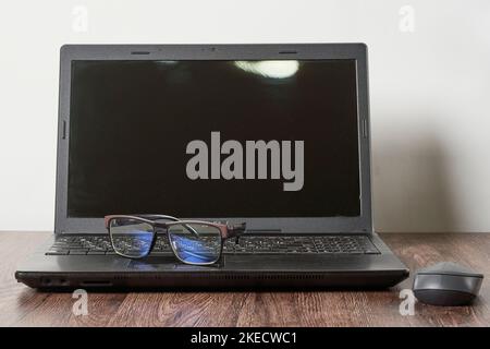 a laptop computer and glasses on a wooden table next to a mouse, with a white wall in the background Stock Photo