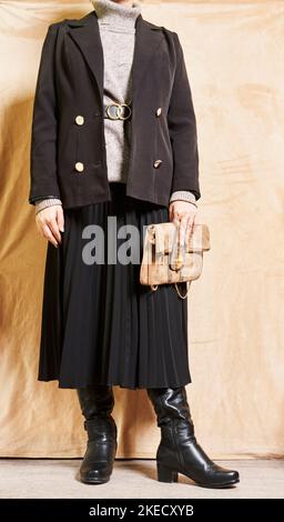 a woman wearing a black coat and holding a brown purse in her right hand, standing against a beige backdrop Stock Photo