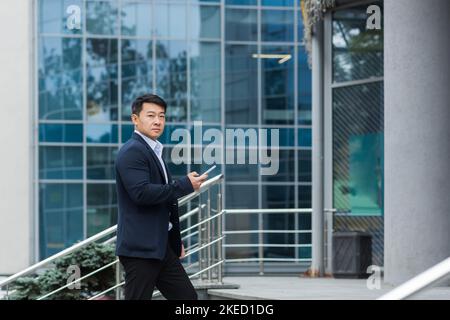 A young handsome Asian man goes to a meeting, an interview for a job in an office center. He is standing on the stairs with a phone and documents in his hands, looking at the camera. Stock Photo