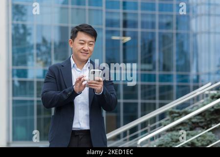 Young handsome male Asian businessman is waiting for a business meeting. He is standing near the office center with a phone in his hands, typing, smiling. Stock Photo
