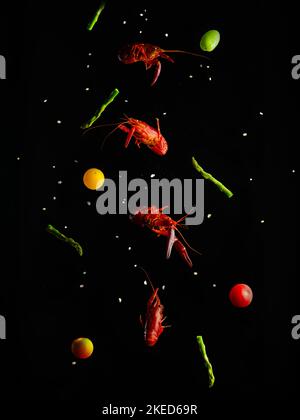 Boiled crayfish, pieces of asparagus, red and yellow cherry tomatoes in a frozen flight on a black background with white grains of salt. Creative culi Stock Photo