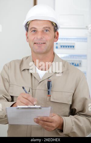 mature electrician with clipboard stood by fusebox Stock Photo