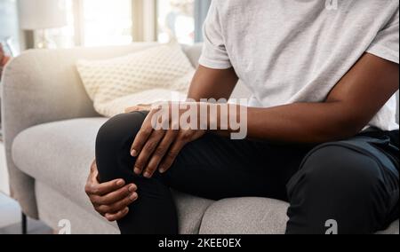 Knee pain, hand of a man and injury while sitting on home living room sofa for rest and relaxation. Ache, painful leg joint and african american man Stock Photo