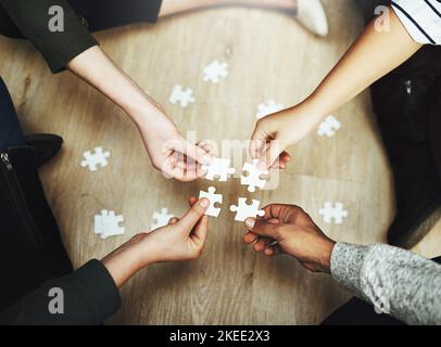 Putting all the pieces together. a group of unrecognizable people fitting puzzle pieces together on the floor. Stock Photo