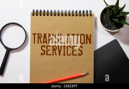 On a light background, gold-framed glasses, a flower in a pot, a green notebook, a red pen and a brown notebook with the text TRANSLATION SERVICES. Bu Stock Photo
