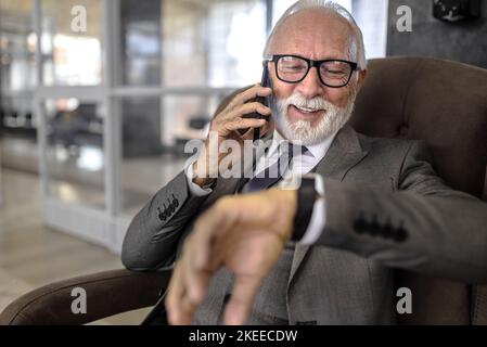 Smiling entrepreneur checking time on wristwatch. Elderly businessman wearing eyeglasses is talking on smart phone. He is waiting at a subway or train Stock Photo
