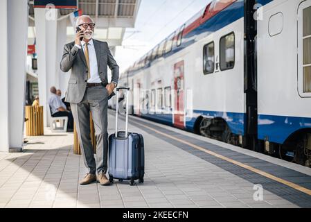 Smiling senior businessman talking on smart phone while standing with hands in pockets. Male entrepreneur is waiting with luggage by train at a railro Stock Photo