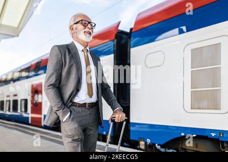 Senior smiling male professional waiting at railroad train station platform. Businessman is standing with luggage by train. He is wearing suit in busi Stock Photo