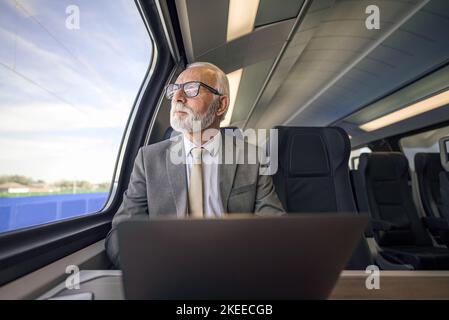 Senior thoughtful businessman with laptop looking through window. Male professional is working while traveling on passenger train. He is wearing eyegl Stock Photo