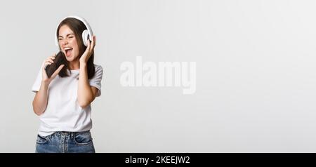 Attractive happy woman playing karaoke app, singing into smartphone, wearing wireless headphones Stock Photo