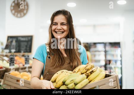 Happy woman working inside supermarket holding a box containing fresh bananas Stock Photo