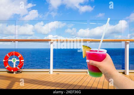 Hand holding a glass with cocktail and straw on a deck of cruise ship with ocean on background with copy space