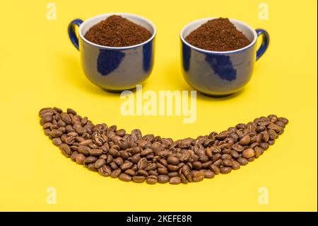 Happy emoticon made of coffee beans on a yellow background, Smiley made of coffee and two cups of ground coffee as eyes. Stock Photo