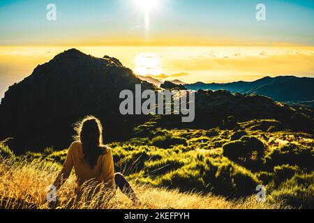 Description: Woman sitting on a mountain meadow enjoying the beautiful mountain landscape of Pico do Ariero at sunrise. Pico do Arieiro, Madeira Islan Stock Photo