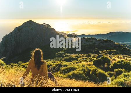 Description: Woman sitting on a mountain meadow enjoying the beautiful mountain landscape of Pico do Ariero at sunrise. Pico do Arieiro, Madeira Islan Stock Photo