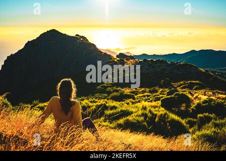 Description: Woman sitting on a mountain meadow enjoying the beautiful mountain landscape of Pico do Ariero at sunrise. Pico do Arieiro, Madeira Islan Stock Photo