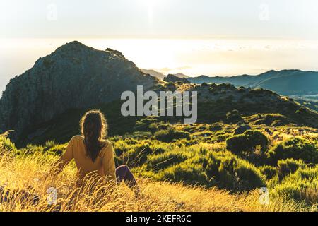 Description: Woman sitting on a mountain meadow enjoying the beautiful mountain landscape of Pico do Ariero at sunrise. Pico do Arieiro, Madeira Islan Stock Photo