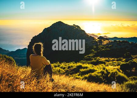 Description: Woman sitting on a mountain meadow enjoying the beautiful mountain landscape of Pico do Ariero at sunrise. Pico do Arieiro, Madeira Islan Stock Photo