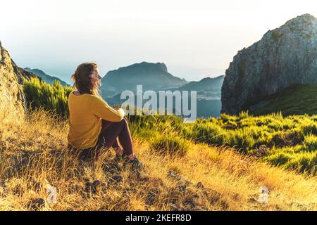 Description: Woman sitting on a mountain meadow enjoying the beautiful mountain landscape of Pico do Ariero at sunrise. Pico do Arieiro, Madeira Islan Stock Photo