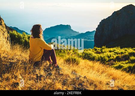 Description: Woman sitting on a mountain meadow enjoying the beautiful mountain landscape of Pico do Ariero at sunrise. Pico do Arieiro, Madeira Islan Stock Photo