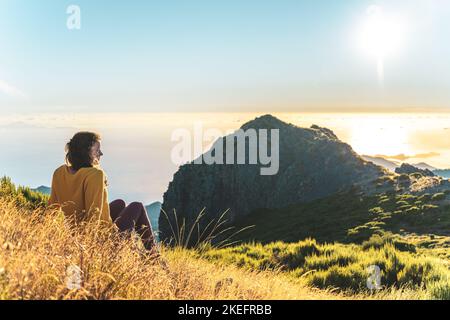 Description: Woman sitting on a mountain meadow enjoying the beautiful mountain landscape of Pico do Ariero at sunrise. Pico do Arieiro, Madeira Islan Stock Photo