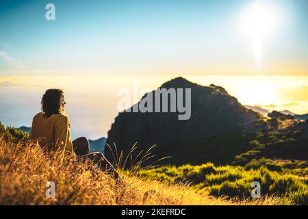 Description: Woman sitting on a mountain meadow enjoying the beautiful mountain landscape of Pico do Ariero at sunrise. Pico do Arieiro, Madeira Islan Stock Photo