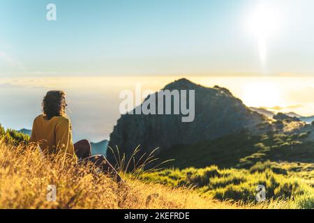 Description: Woman sitting on a mountain meadow enjoying the beautiful mountain landscape of Pico do Ariero at sunrise. Pico do Arieiro, Madeira Islan Stock Photo