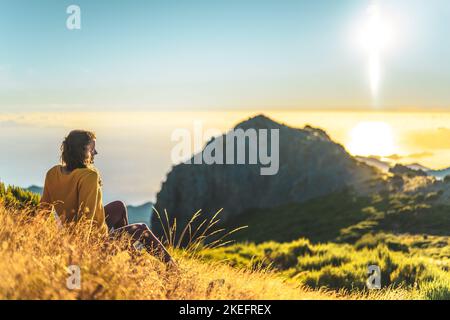 Description: Woman sitting on a mountain meadow enjoying the beautiful mountain landscape of Pico do Ariero at sunrise. Pico do Arieiro, Madeira Islan Stock Photo