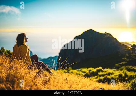 Description: Woman sitting on a mountain meadow enjoying the beautiful mountain landscape of Pico do Ariero at sunrise. Pico do Arieiro, Madeira Islan Stock Photo