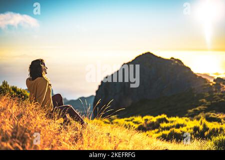 Description: Woman sitting on a mountain meadow enjoying the beautiful mountain landscape of Pico do Ariero at sunrise. Pico do Arieiro, Madeira Islan Stock Photo