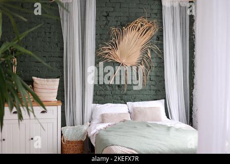 Modern boho interior of bedroom with green brick wall, bed, white wooden commode, curtains, houseplants, rattan basket, dray palm leaf and design pers Stock Photo