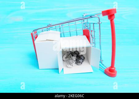 Miniature steel dumbbells in a small shipping box placed in an overturned toy shopping trolley isolated on a blue wooden background. Buying gym equipm Stock Photo