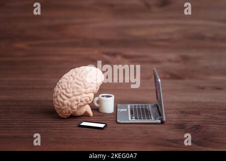 Human brain figurine placed on a wooden table with toy laptop, smartphone and coffee cup. Creative busy brain concept. Stock Photo