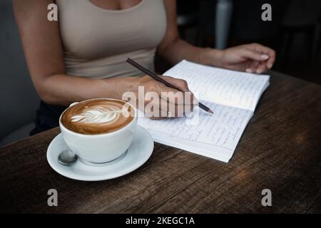 Unrecognizable woman sits at table in cafe with cup of coffee and writes in notebook notes writing information Stock Photo
