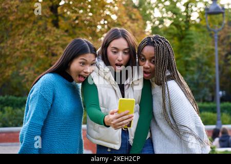 Three multiethnic female ladies amazed watching cellphone, standing outside at city public park. Stock Photo