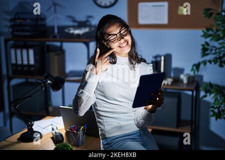 Young brazilian woman using touchpad at night working at the office smiling cheerful showing and pointing with fingers teeth and mouth. dental health Stock Photo