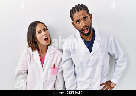 Young hispanic doctors standing over white background in shock face, looking skeptical and sarcastic, surprised with open mouth Stock Photo