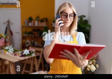 Young blonde woman florist talking on smartphone reading book at flower shop Stock Photo