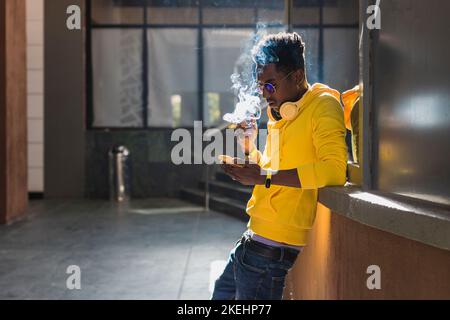 A young black adult male dressed in a yellow sweatshirt and blue pants is smoking leaning against a wall while looking at his cell phone screen. Stock Photo