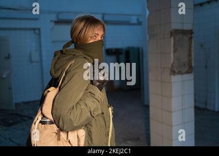 woman in a mask and with a backpack walks through an old abandoned large building. concept of a survivor wandering in the post-apocalyptic world in Stock Photo